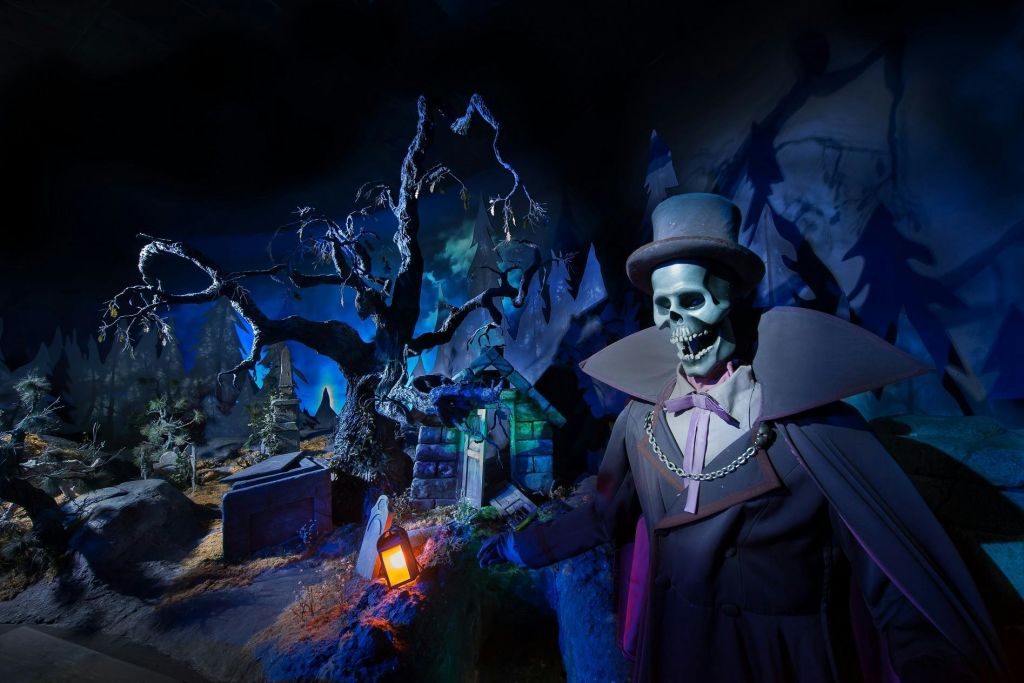 상단 모자와 망토를 두른 해골은 뒤틀린 나무, 비석, 등불이 있는 어둡고 소름끼치는 곳에 서 있습니다.