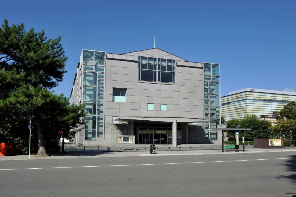 커다란 창문과 중앙 출입구, 맑은 하늘 배경이 돋보이는 현대적인 회색 건물입니다. 측면에 나무가 부분적으로 보입니다.