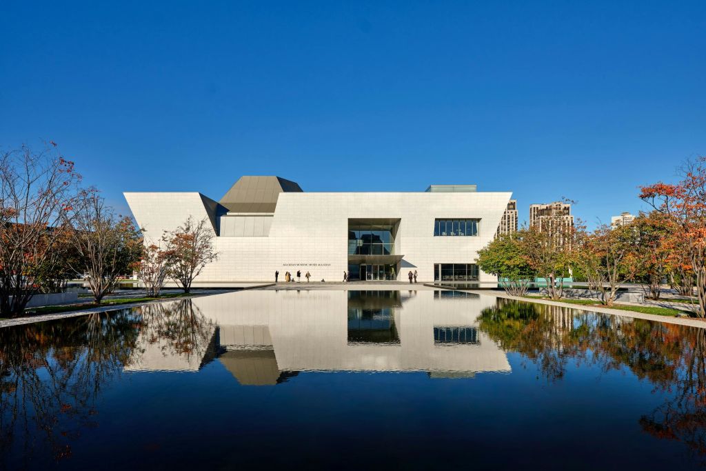 청명한 푸른 하늘 아래 고요한 수영장에 반사된 각진 건축물이 있는 현대적인 흰색 건물입니다.