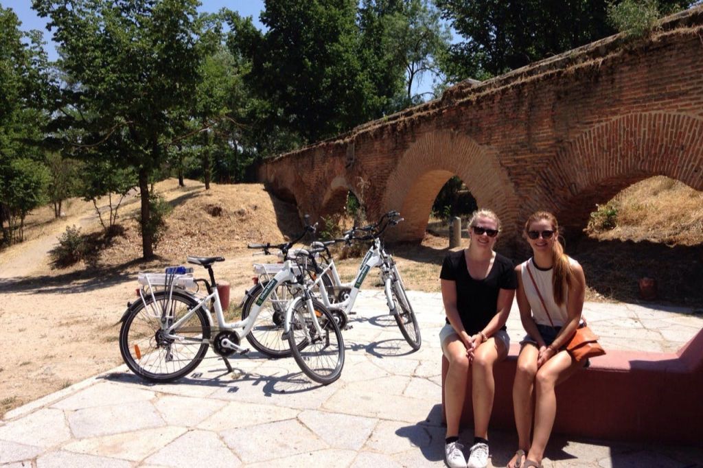 나무로 둘러싸인 인근에 세 대의 자전거가 주차된 벽돌 아치형 다리 앞 벤치에 두 사람이 앉아 있습니다.