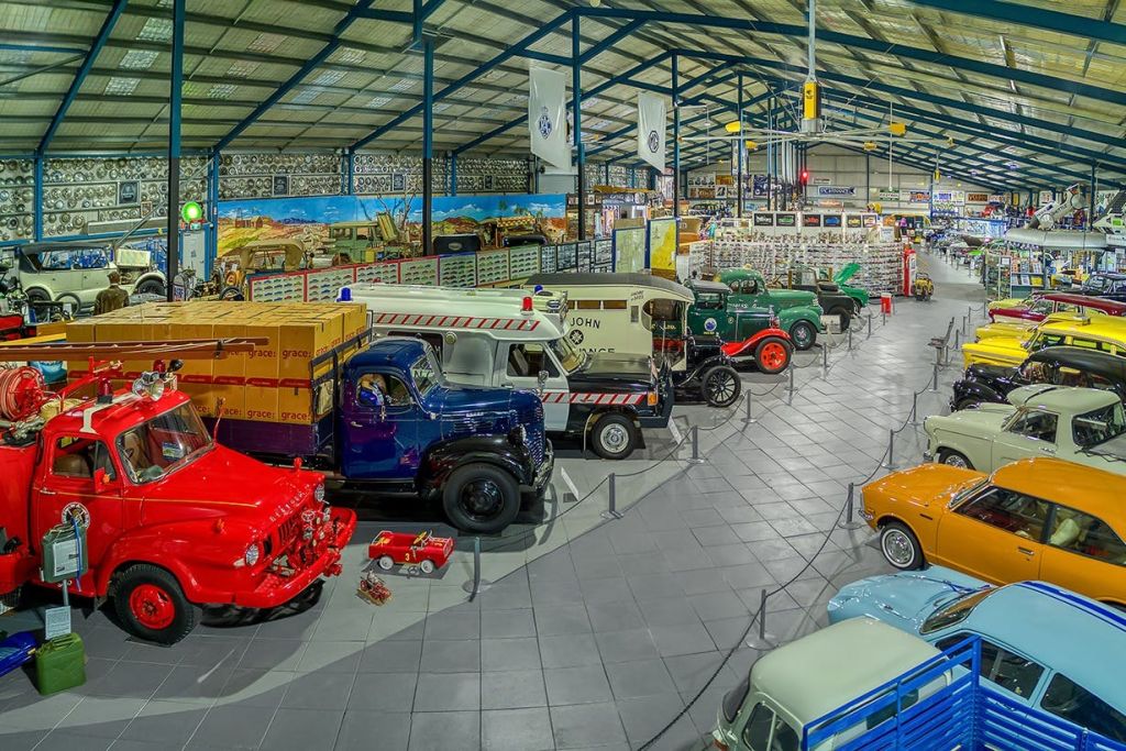 조명이 밝은 대형 홀 내부의 타일 바닥에 다양한 색상의 빈티지 트럭과 밴이 줄지어 전시된 박물관 전시관입니다.