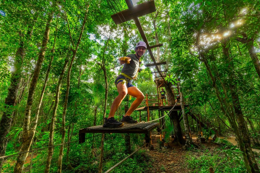 숲속 어드벤처 파크의 밧줄 다리에서 울창한 녹음으로 둘러싸인 헬멧과 하네스를 착용하고 균형을 잡는 사람.