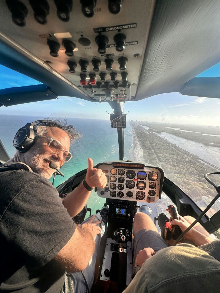 헤드셋을 착용한 사람이 해안선을 가로지르는 헬리콥터 조종석 안에서 엄지손가락을 치켜세우고 있습니다.