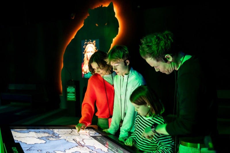 한 무리의 사람들이 조명이 어두운 방에서 터치 스크린 디스플레이와 상호 작용하며, 배경에는 빛나는 늑대 실루엣이 있습니다.