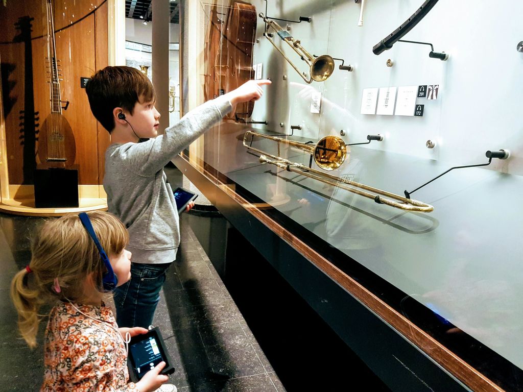 헤드폰을 든 두 어린이가 박물관 케이스에 전시된 악기를 보고 있다. 하나는 트럼펫을 가리키고 있다.