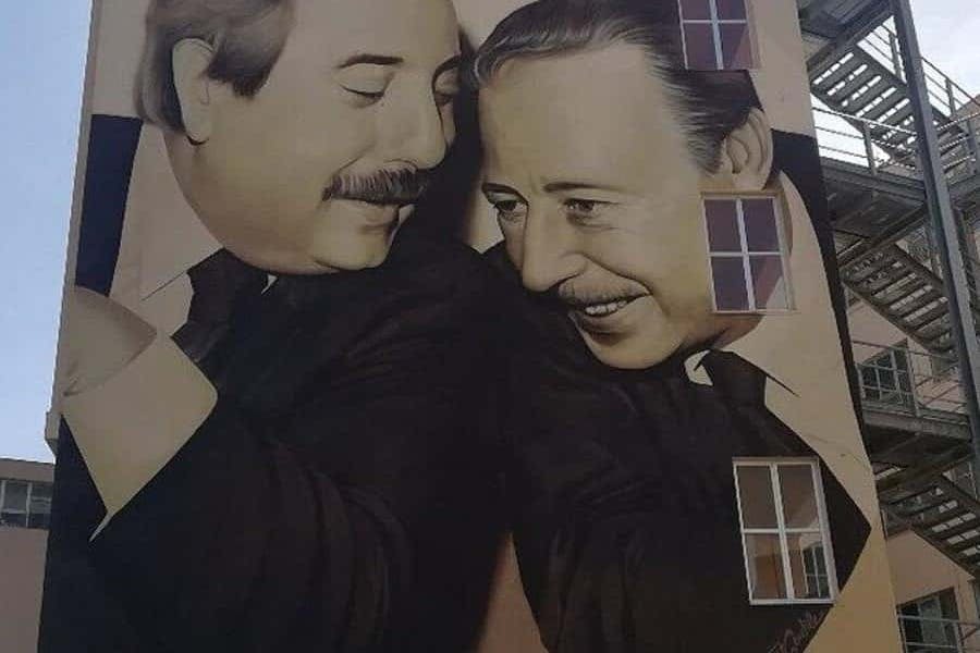 콧수염을 한 두 남자가 미소를 지으며 벽화에서 서로를 마주보고 있습니다. 벽화에는 창문이 걸려 있습니다.