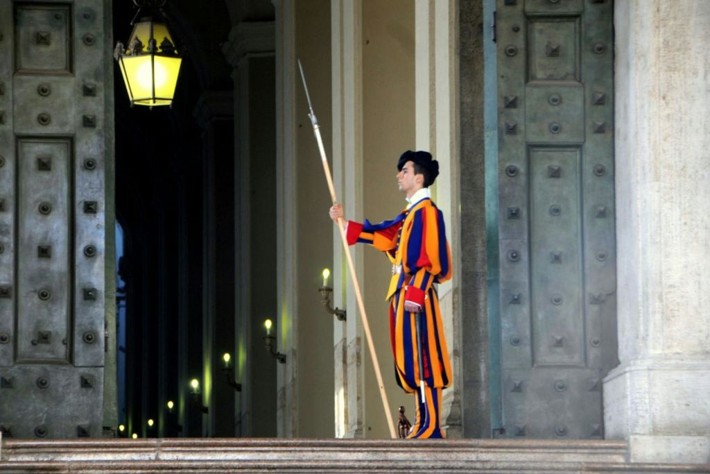 긴 창을 들고 있는 화려한 유니폼을 입은 경비원이 장식 패널과 여러 개의 조명 벽 램프가 있는 웅장한 출입구에 서 있습니다.