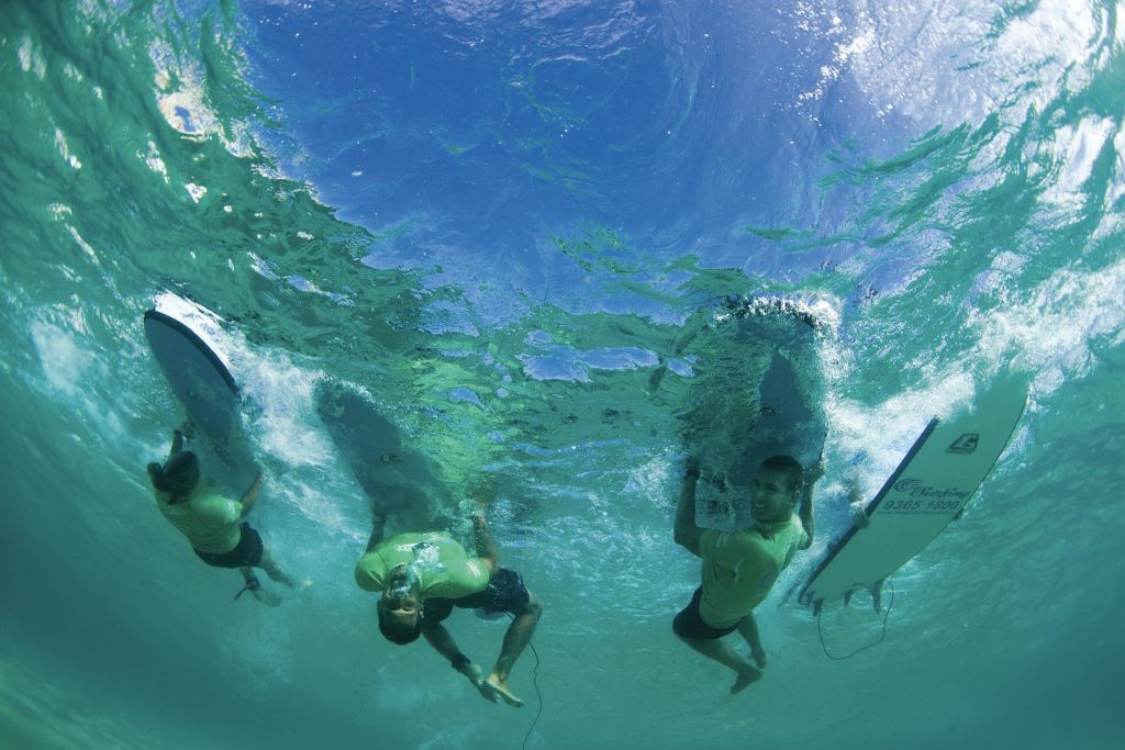 녹색 셔츠를 입은 세 명의 서퍼가 수중 서핑보드에서 노를 젓고 있으며, 수면 위로 하늘이 보입니다.