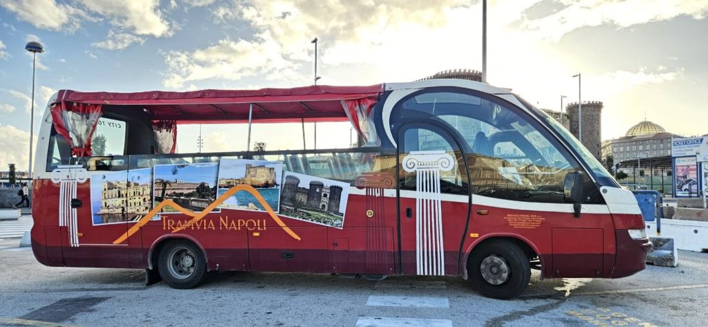 화창한 날에 주차된 성곽과 기둥을 포함한 나폴리 랜드마크의 이미지가 있는 빨간색 관광 버스.
