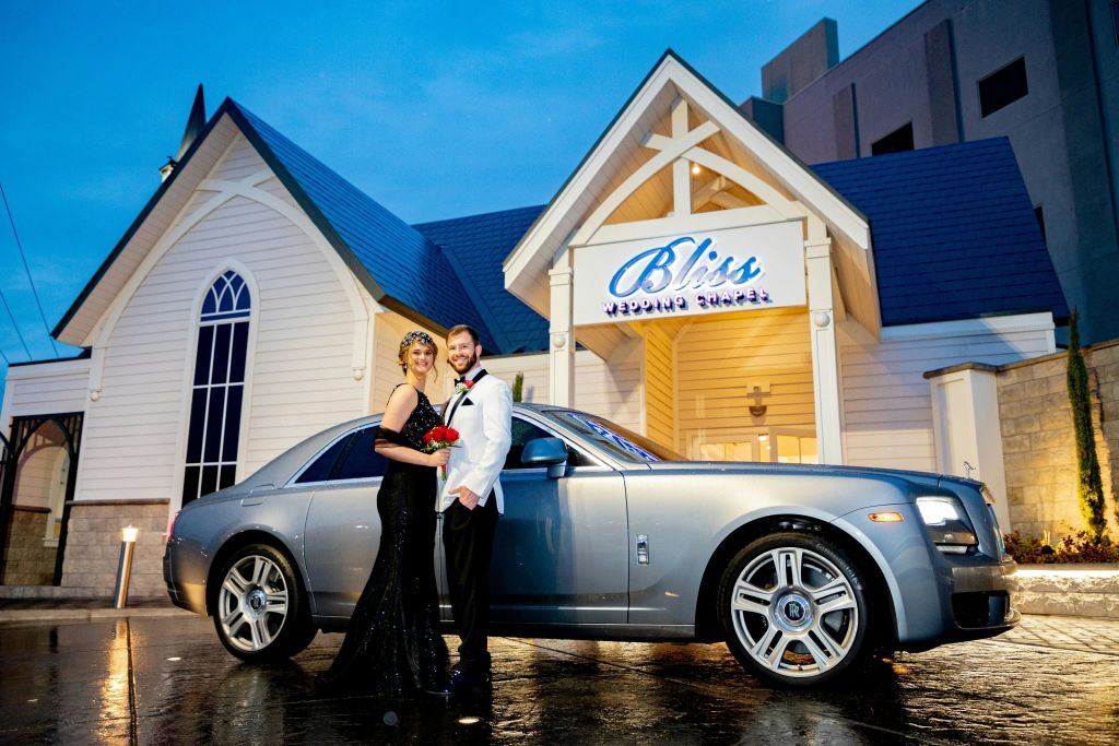 "Bliss Wedding Chapel" 표지판이 있는 건물 밖에서 정장 차림으로 웃고 있는 커플이 고급 승용차 옆에 서 있습니다.