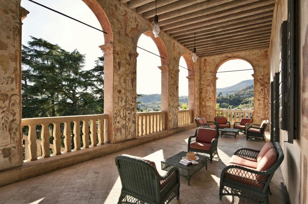 장식된 아치, 고리버들 의자, 나무와 언덕이 내려다보이는 책이 놓인 테이블이 있는 야외 발코니가 있습니다.