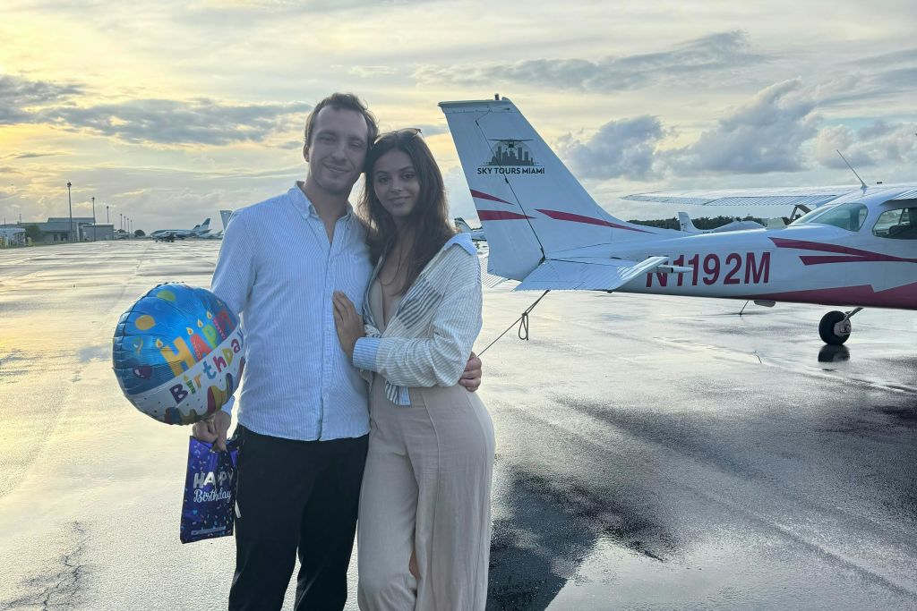 부부가 작은 비행기 근처의 젖은 활주로에 서 있습니다. 남자는 미소를 지으며 생일 풍선과 선물 가방을 들고 있습니다.