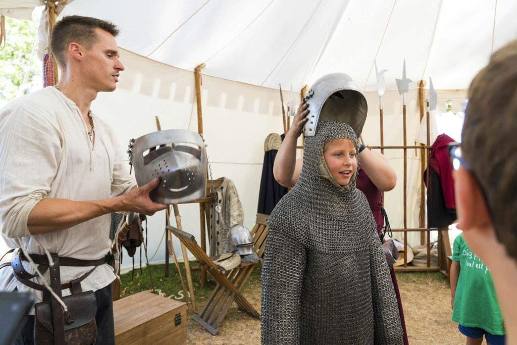 어린이가 텐트 안에서 중세 기사의 헬멧을 착용하고 갑옷과 중세 물품을 전시하는 것을 어른이 도와줍니다.