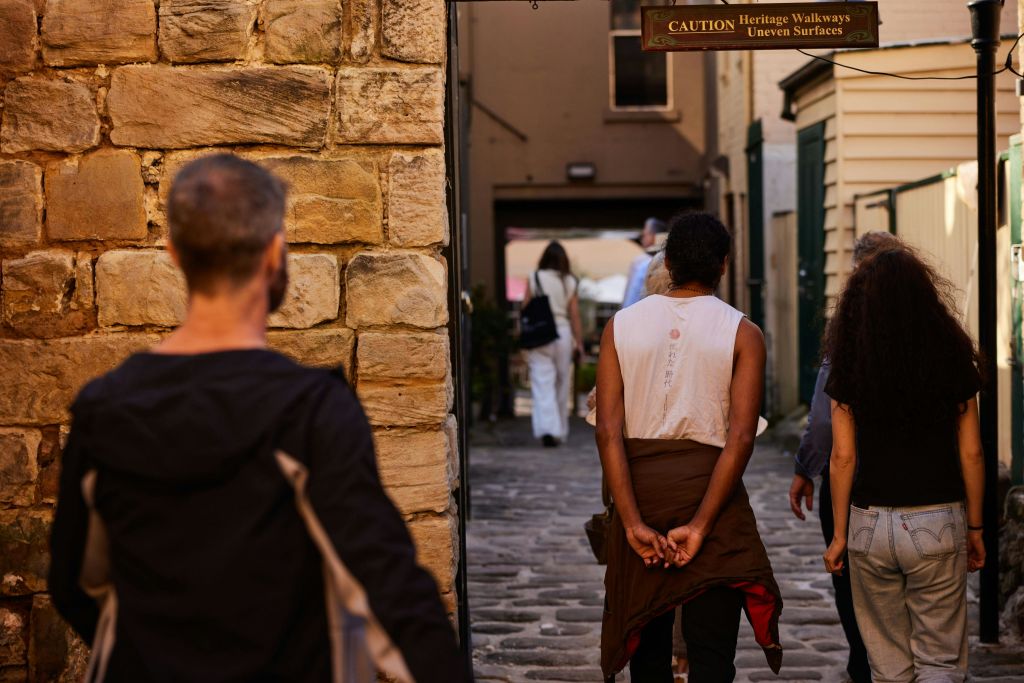 유서 깊은 성벽이 있는 좁은 돌로 포장된 골목을 거닐고 있는 사람들, 위에는 주의 표지판이, 뒤에는 건물이 있습니다.