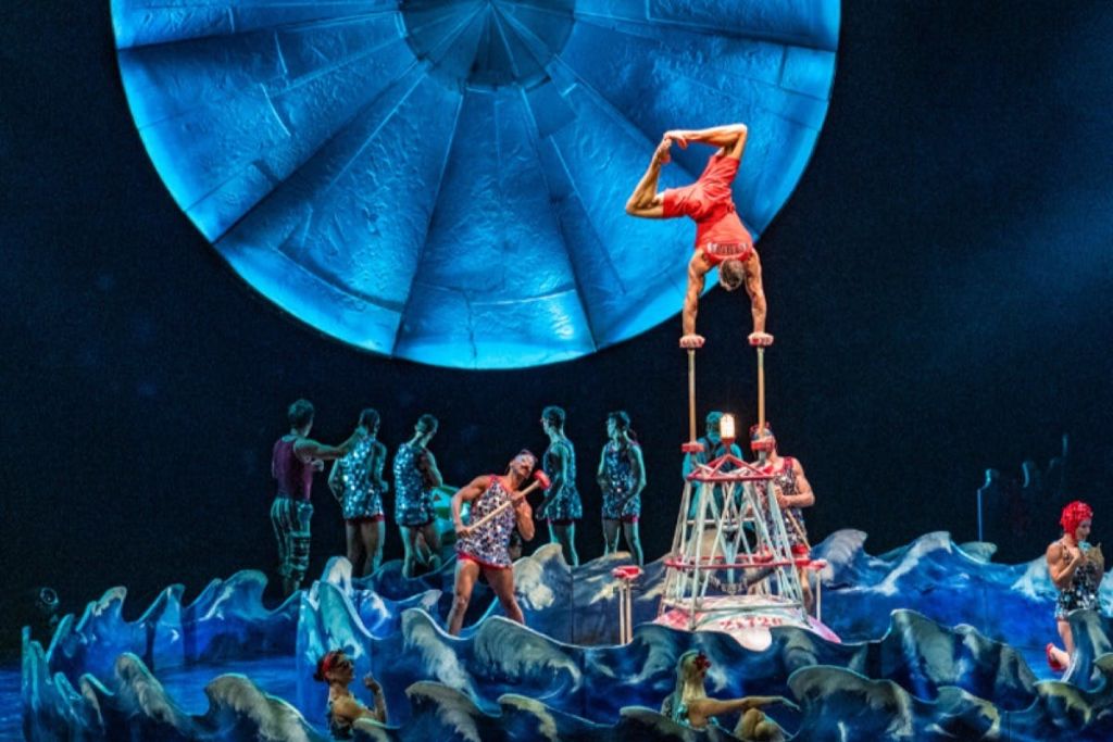 아크로바트는 파란색 돔 아래에서 다른 연주자와 소품으로 둘러싸인 구조물 위에서 손에 균형을 잡습니다.