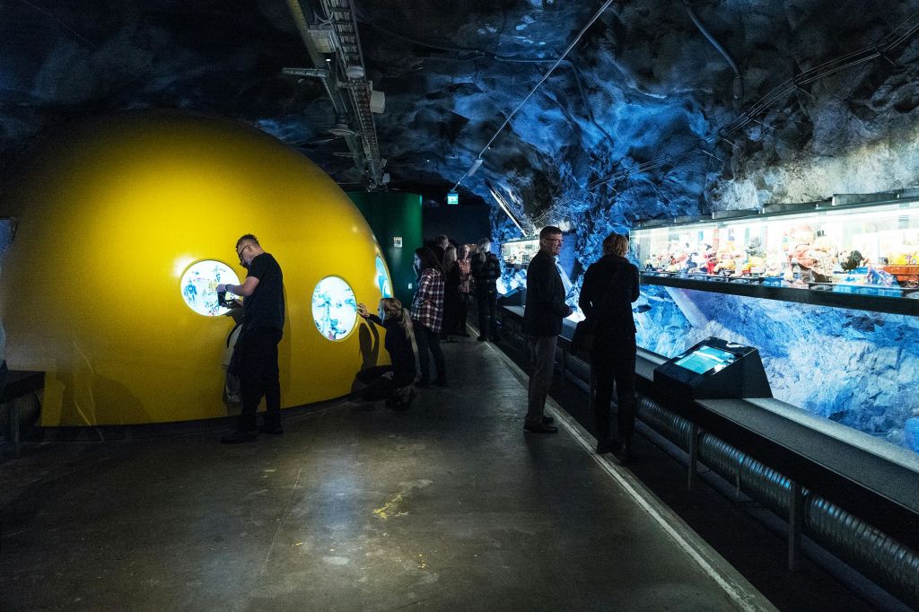 조명이 어두운 터널 같은 방 안의 커다란 노란색 돔 모양의 구조물에서 창문을 통해 전시물을 관찰하는 사람들.