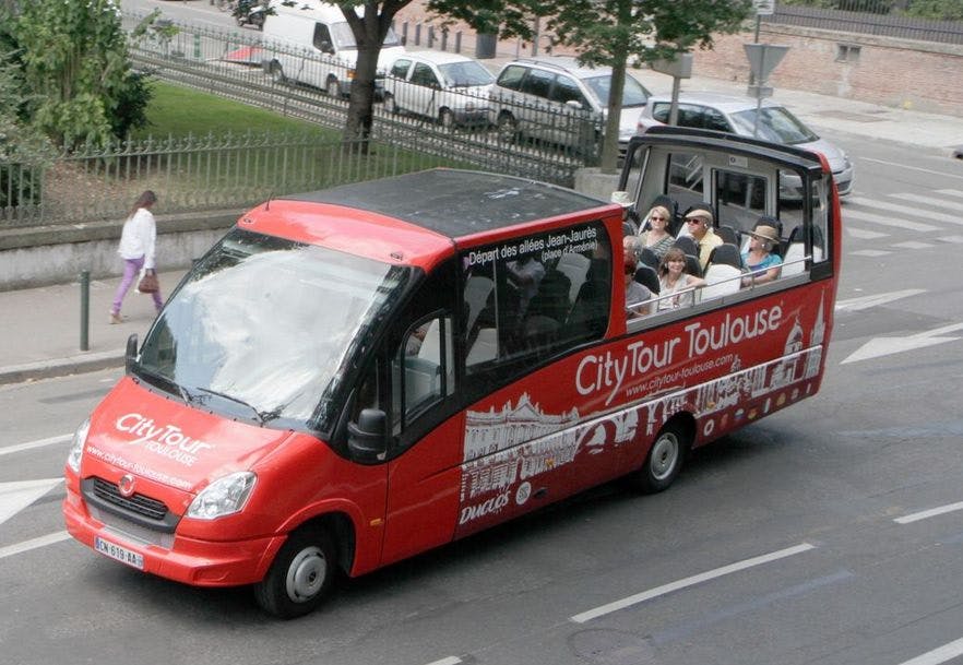 빨간색 시티 투어 툴루즈 버스가 탑승객들과 함께 탁 트인 갑판 위에 앉아 도심 거리를 달립니다.