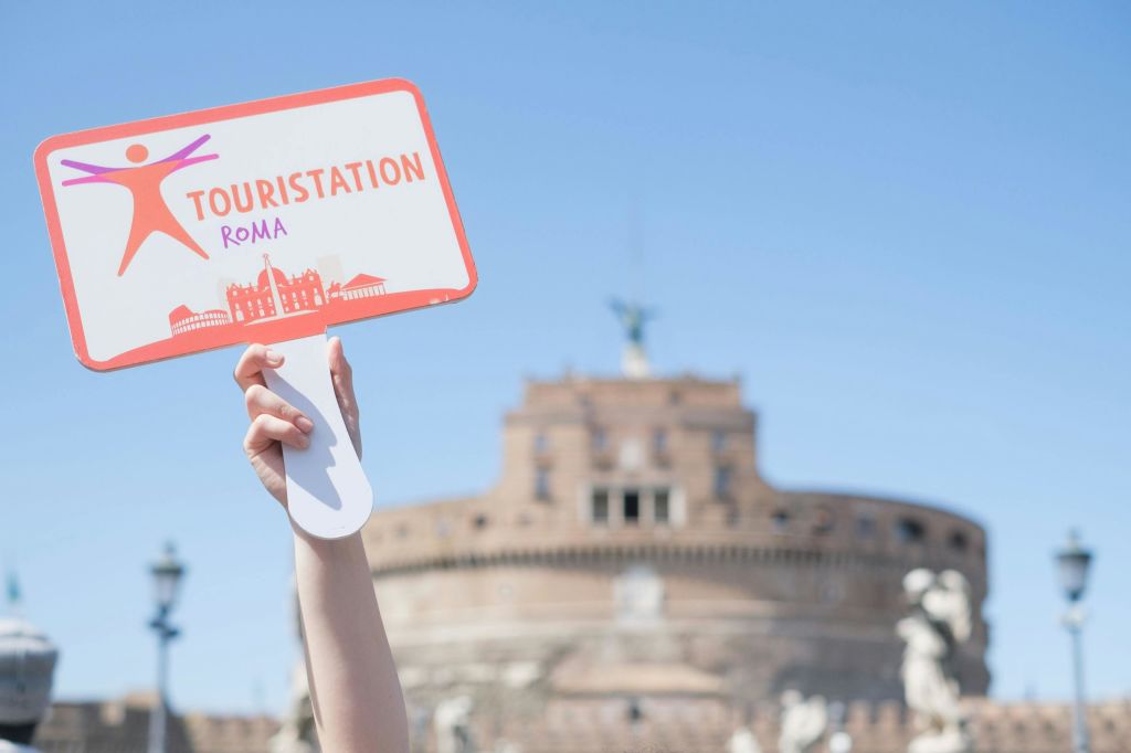 파란 하늘을 배경으로 산탄젤로 성 앞에서 'Touristation Roma' 라고 적힌 표지판을 들고 있는 손.