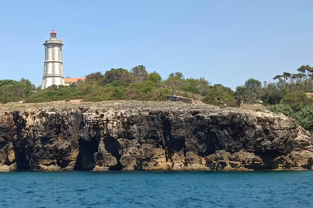 맑고 푸른 하늘 아래 물에서 본 하얀 등대와 초목이 우거진 바위 해안선.