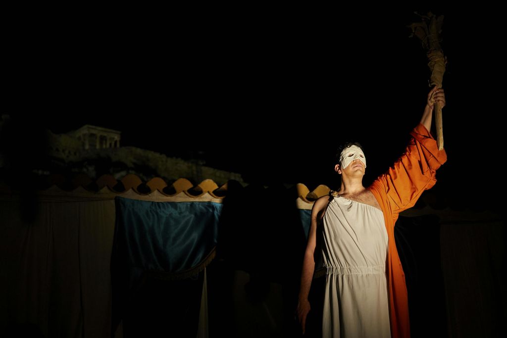 흰색 토가와 주황색 망토를 입은 사람이 흰색 마스크를 쓰고 어두운 배경에 횃불을 들고 있습니다.