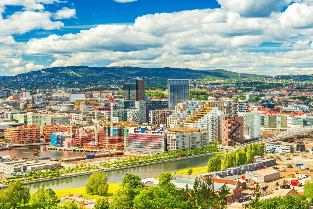 먼 언덕이 있는 부분적으로 흐린 하늘 아래 현대적인 건물, 강, 주변 녹지가 있는 다채로운 도시 풍경.