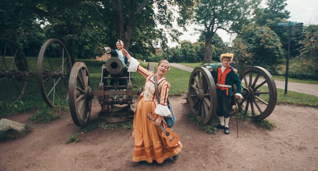 공원의 대포 근처에서 역사적인 의상을 입은 세 사람. 하나는 꼭두각시, 다른 하나는 우쿨렐레, 다른 하나는 대포 옆에 서 있습니다.