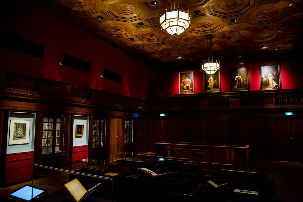 벽에 나무 패널, 유리 케이스의 책, 샹들리에, 액자에 담긴 초상화가 있는 조명이 희미하고 화려한 서재입니다.