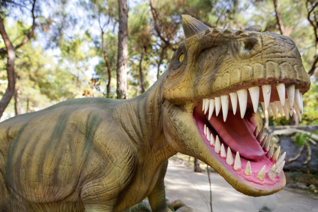 나무와 산책로를 배경으로 날카로운 이빨을 드러내는 입을 벌린 공룡 동상의 클로즈업.