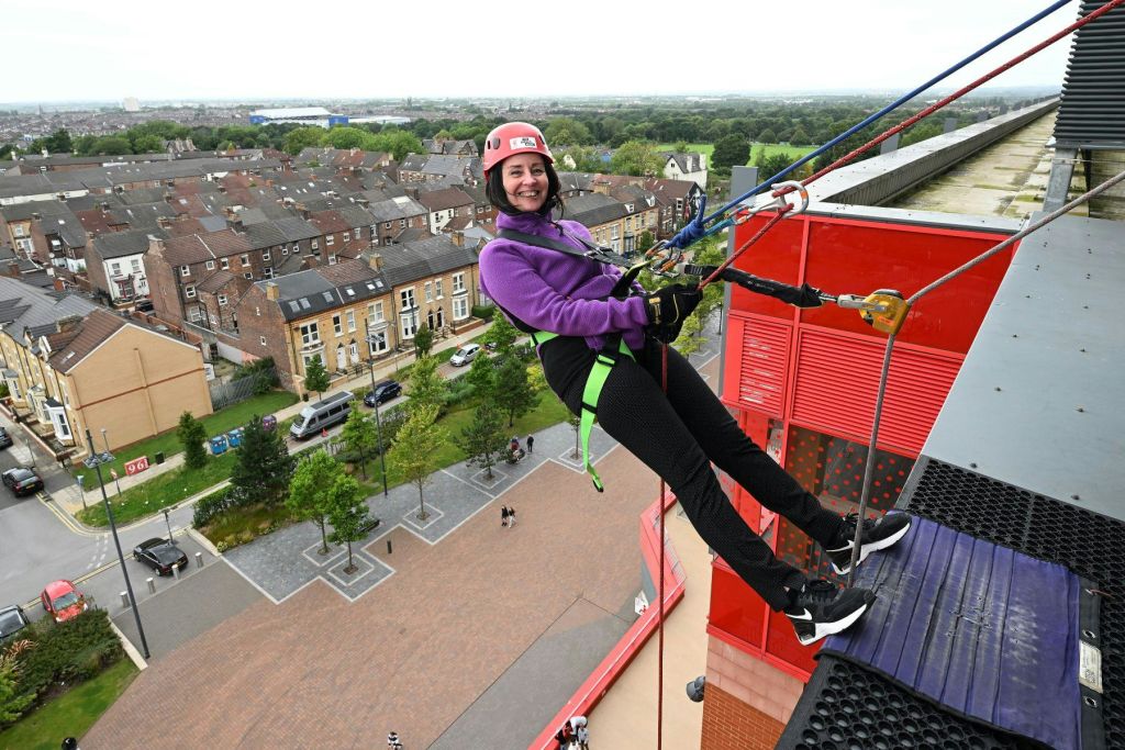 헬멧을 쓰고 하네스를 착용한 여성이 빨간색 건물을 내려가며 미소를 짓고 있습니다. 주택가와 녹지를 배경으로 말이죠.