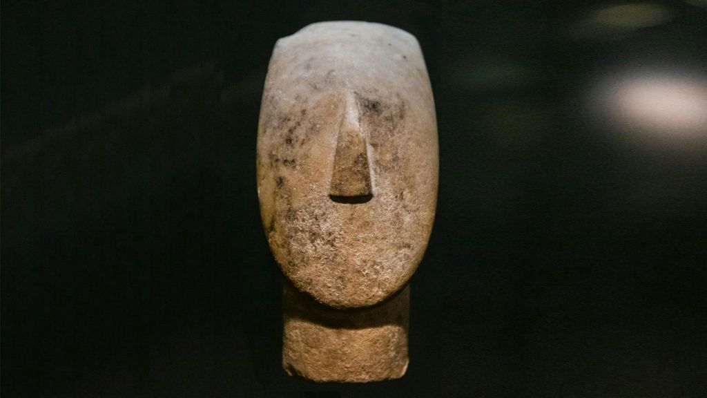 어두운 배경에 심플하고 추상적인 얼굴 디자인이 돋보이는 양식화된 인간 머리의 석조 조각.