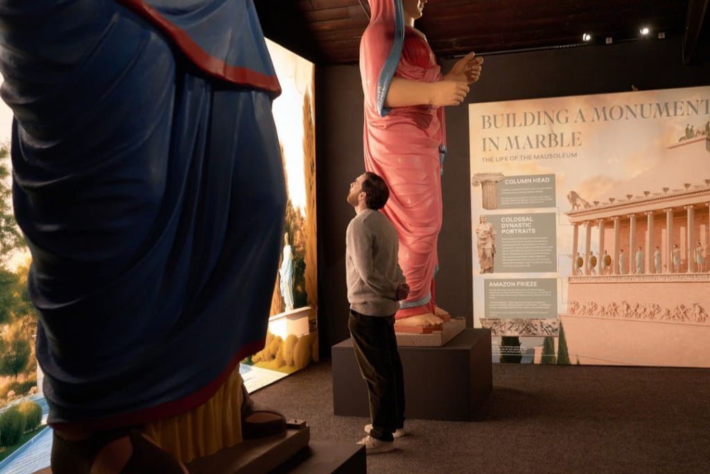 한 사람이 대형 다채로운 조각상과 고대 건축물에 대한 정보 패널이 있는 박물관 전시물을 관찰합니다.