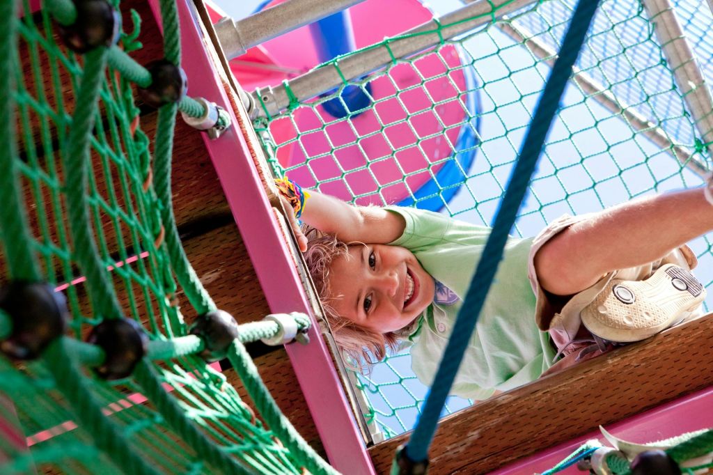 화창한 하늘 아래 밧줄과 그물로 둘러싸인 다채로운 놀이 구조물을 보며 미소 짓고 등반하는 아이.