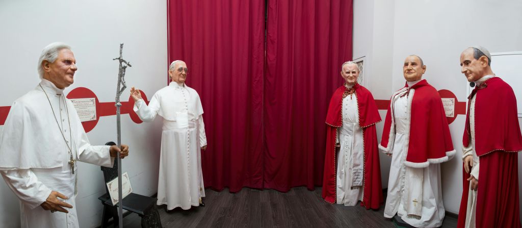 빨간 망토와 흰색 가운을 입은 실물 크기의 남성 조각상 세 개가 빨간색 커튼과 흰색 벽에 전시되어 있습니다.