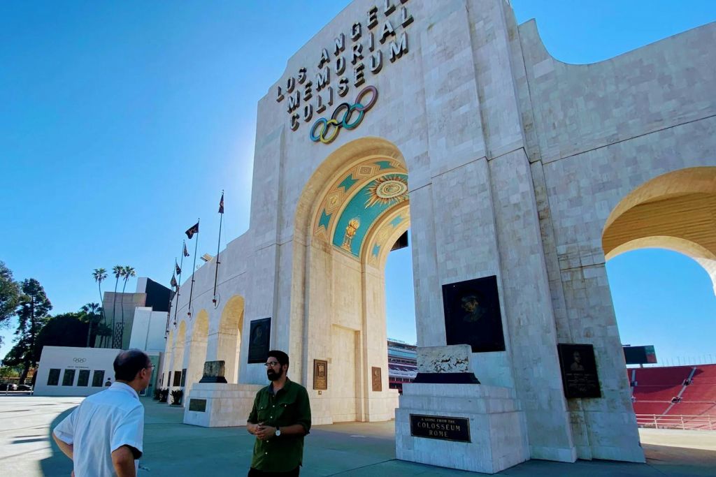 맑은 날, 올림픽 고리와 커다란 아치형 통로가 있는 로스앤젤레스 메모리얼 콜리세움 밖에 두 사람이 서 있습니다.