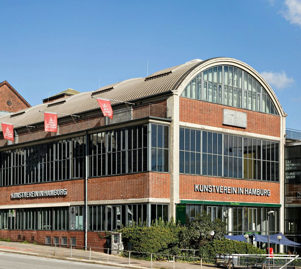 "Kunstverein in Hamburg" 라고 쓰여진 대형 벽돌과 유리 건물에는 빨간색 배너와 곡선형 지붕이 있습니다.