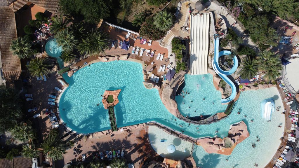 초목과 통로로 둘러싸인 수영장, 미끄럼틀, 라운지 의자가 있는 워터파크의 공중 전망.