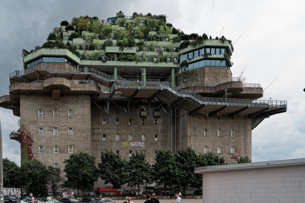 옥상에 녹색 테라스와 나무가 있는 대형 콘크리트 건물로 여러 개의 창문과 앞을 걷는 사람들이 있습니다.