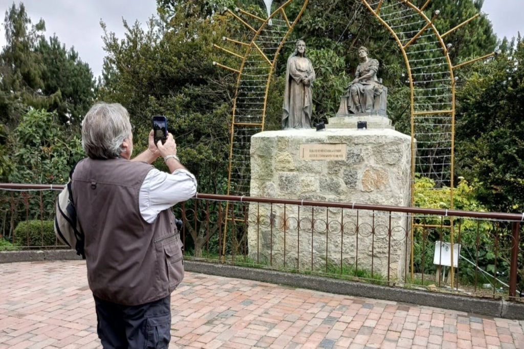 한 사람이 녹지와 금속 아치로 둘러싸인 공원에서 두 개의 석상이 있는 돌 받침대를 촬영하고 있습니다.
