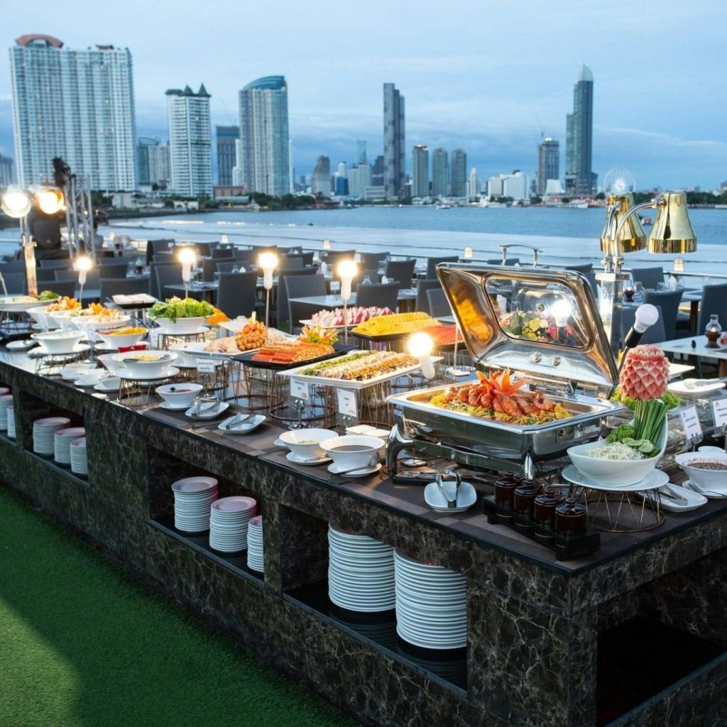 해질 무렵 물가의 도시 스카이라인을 배경으로 다양한 요리와 디저트를 즐길 수 있는 야외 뷔페 시설입니다.