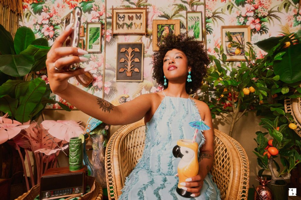 파란색 드레스를 입은 사람이 고리버들 의자에 앉아 화려한 장식과 녹색 식물로 둘러싸인 열대 음료를 들고 있습니다.
