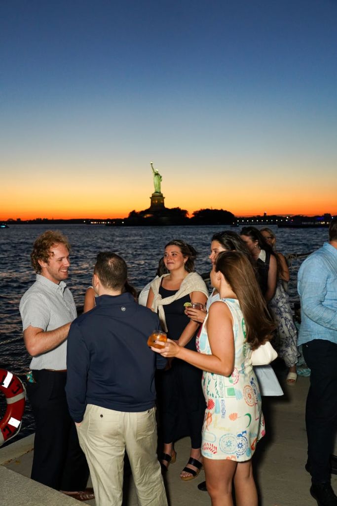 자유의 여신상을 배경으로 노을이 질 무렵 물 근처에서 야외 사교 활동을 하는 사람들.