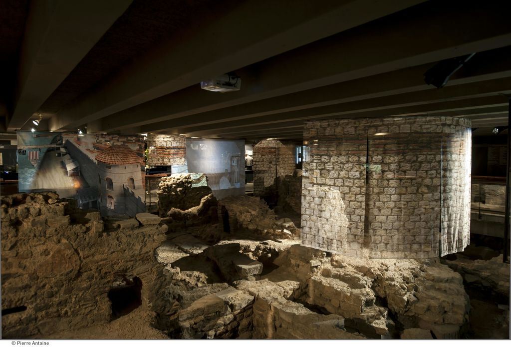 벽돌 벽과 나무 기둥이 있는 건물 아래에 있는 고대 석조 유적에는 벽에 투영된 오래된 구조물의 기둥과 이미지가 있습니다.