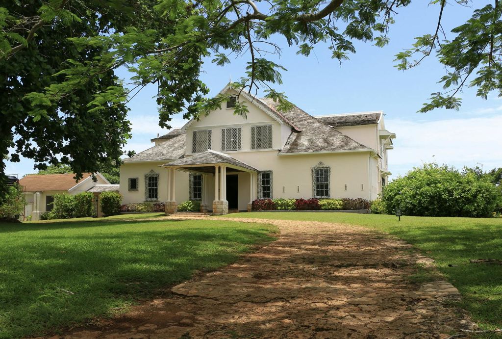 푸른 하늘 아래 푸른 녹음과 나무로 둘러싸인 현관이 있는 크림색의 대형 주택입니다.