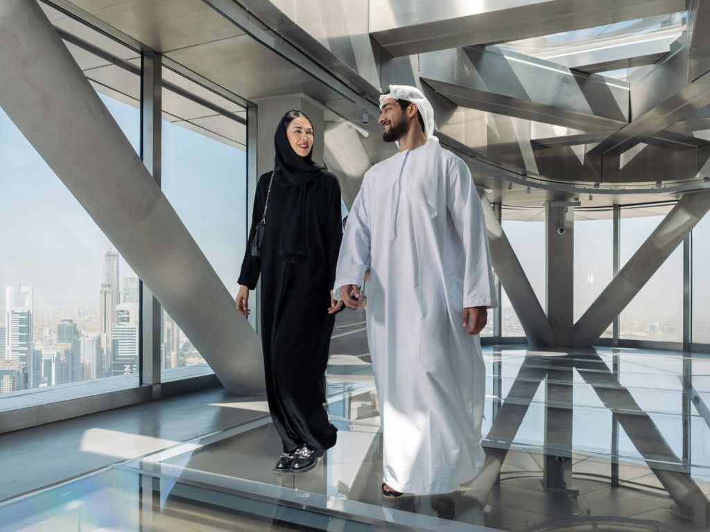 중동 전통 복장을 한 남녀가 도시 전망을 배경으로 한 현대적인 유리 건물에서 나란히 걸어가고 있습니다.