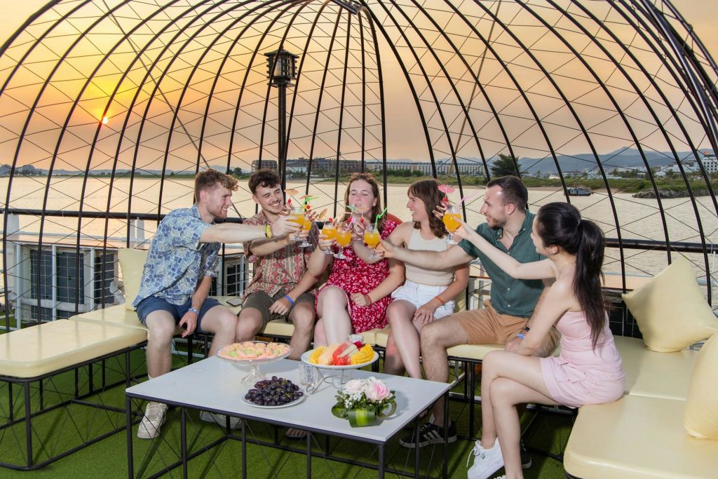 6명의 일행이 정자에 앉아 다채로운 음료와 함께 건배합니다. 앞에 간식과 꽃이 놓인 테이블이 있습니다.