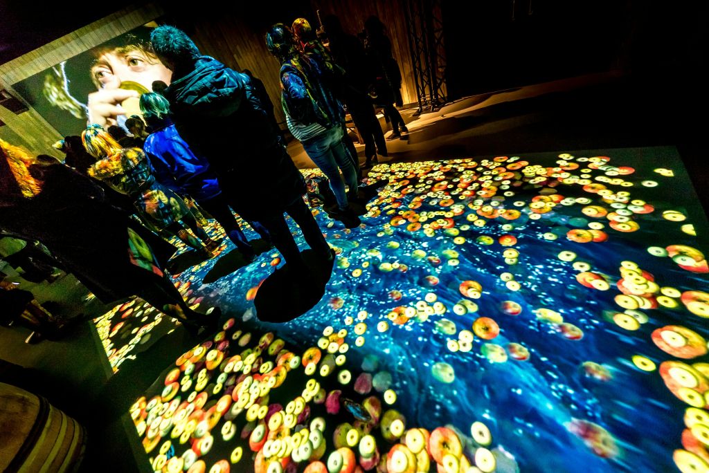 사람들은 바닥에 서서 커다란 얼굴과 추상적인 패턴을 포함하여 화려하고 투영된 예술적 이미지가 있는 벽으로 둘러싸여 있습니다.