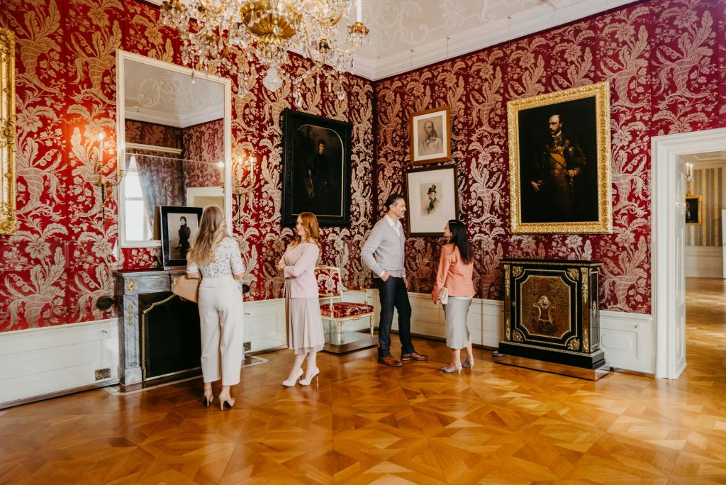 빨간색 패턴의 벽과 액자에 담긴 그림이 있는 호화로운 방에서 네 명이 예술을 감상합니다. 샹들리에 오버헤드와 화려한 가구가 있습니다.