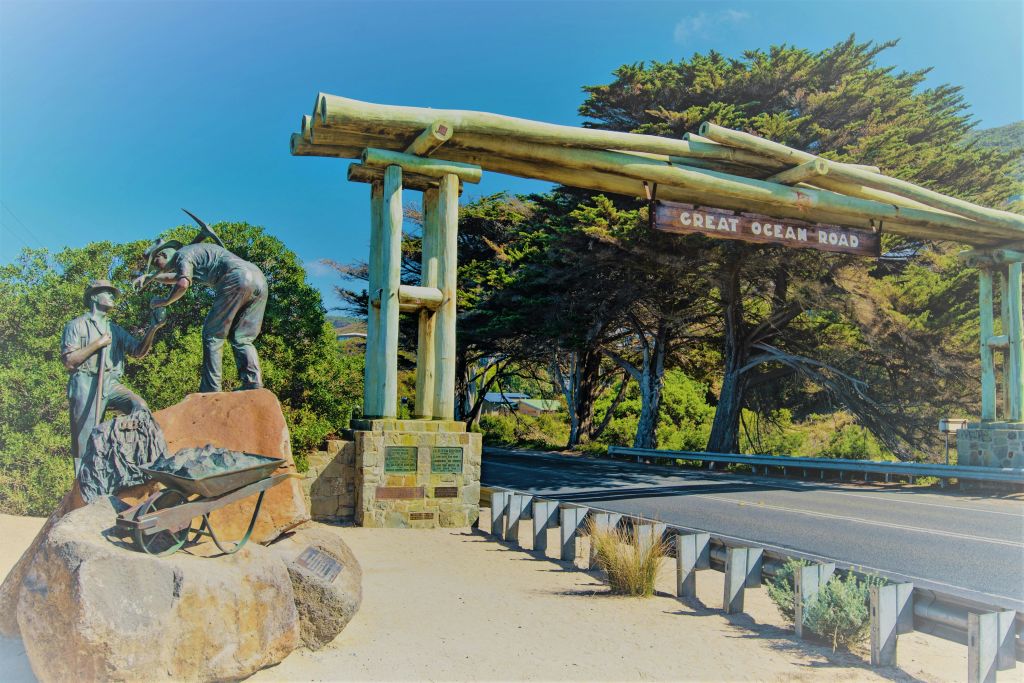 나무로 둘러싸인 손수레를 든 작업자의 조각상 옆에 '그레이트 오션 로드' 라고 쓰인 석조 아치형 통로가 있습니다.