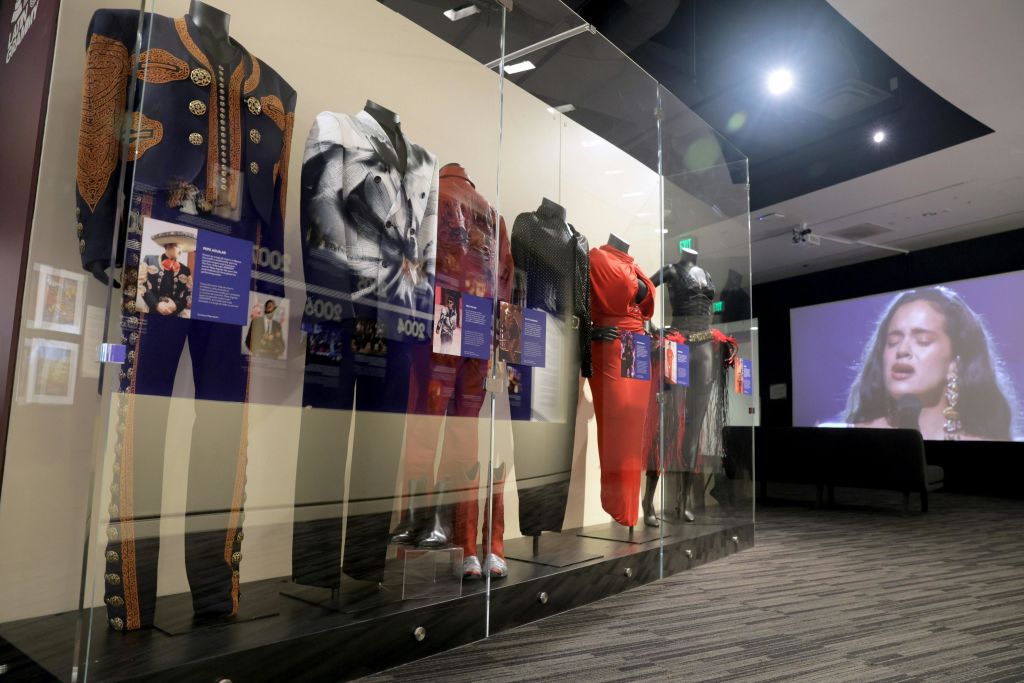 유리 뒤에 다양한 의상을 입은 마네킹이 전시된 박물관 전시관에는 한 여성이 노래하는 대형 스크린이 있습니다.
