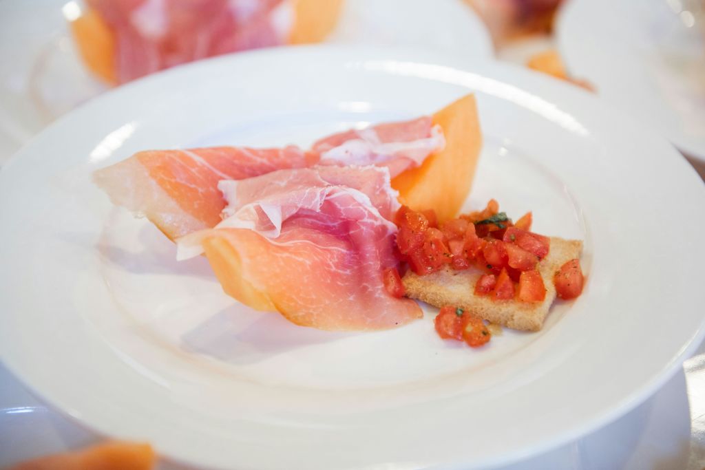 멜론 위에 얇게 썬 프로슈토와 잘게 썬 토마토를 얹은 토스트 조각이 담긴 흰색 접시.